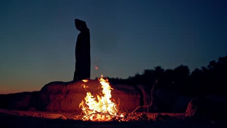 Scary view of an evil in black costume at night near fire. Silhouette of death with hood turning around outdoors. Halloween evening.