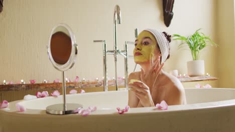 Biracial-woman-in-bath-looking-into-mirror-and-applying-face-mask