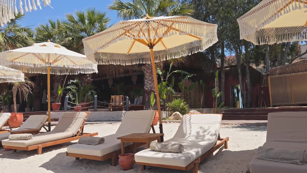 Inquadratura statica grandangolare di lettini di lusso e ombrelloni boho chic con frange allineati sulla sabbia bianca in un esclusivo resort sulla spiaggia tropicale sotto un cielo azzurro e limpido.
