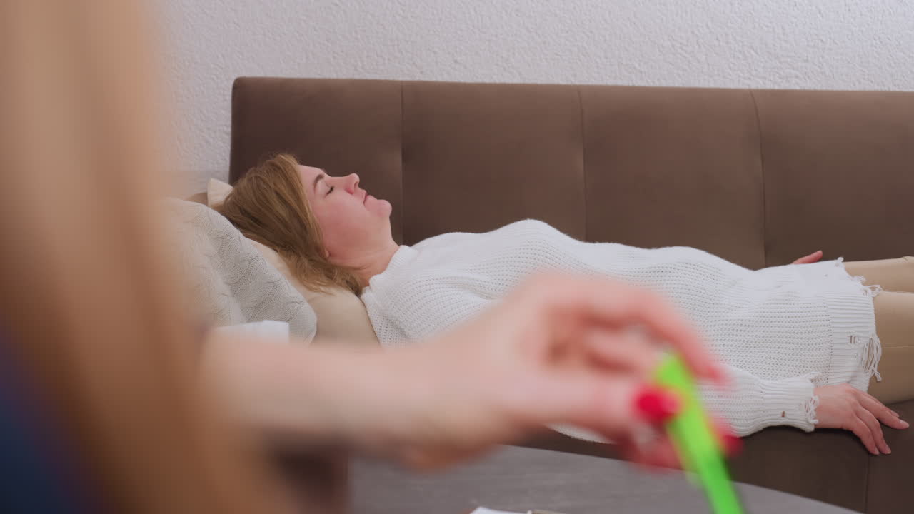 Healing companion reclines on comfortable sofa with eyes closed mid speech during calm conversation with reflective listener holding green pen in serene therapeutic space fostering emotional openness