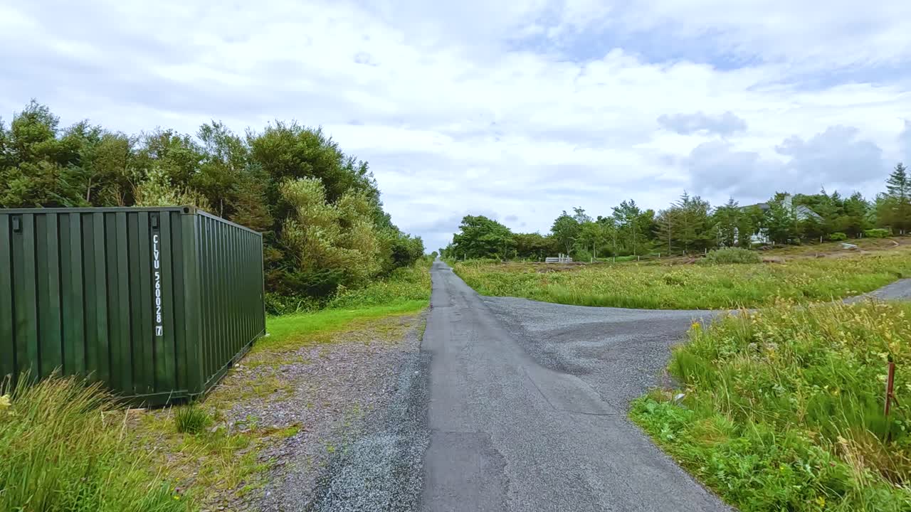 A vehicle travels along a quiet, paved country road bordered by green fields, trees, and a metal storage container under bright natural daylight