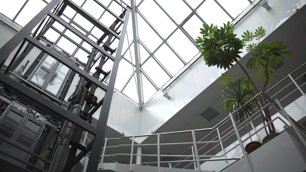 Vista de baixo ângulo dentro de um átrio multinível olhando para cima em direção a uma claraboia de vidro triangular, montantes de aço que se cruzam, estrutura de elevador, grades de varanda, linhas de escada e plantas em