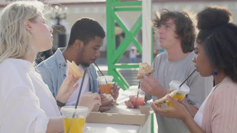 grupo de amigos compartiendo una pizza y bebiendo bebidas frías parados en una mesa al aire libre en la calle, mientras conversan juntos