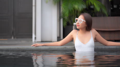 원피스 수영복을 입은 젊은 아시아 여성이 야외 수영장 가장자리에 팔을 기대고 슬로우 모션
