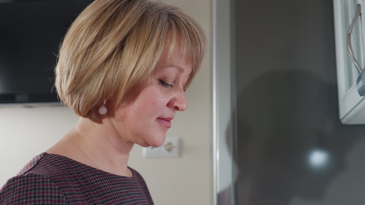 Elegant woman in plaid clothing wearing beautiful earrings dances while focusing on something, partial view of kitchen and TV screen in background