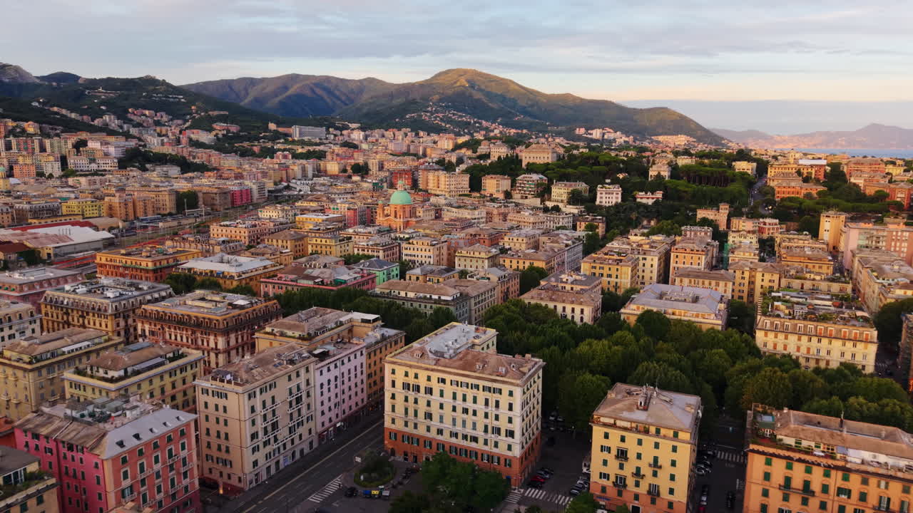 Drone moves forward above Genoa’s rooftops showing colorful buildings, churches, trees, streets, sea in the background and mountains at sunset