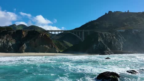 Cinematic-shot-of-Rocky-Creek-Bridge-as-blue-waves-crash-on-shore,-Highway-1-California