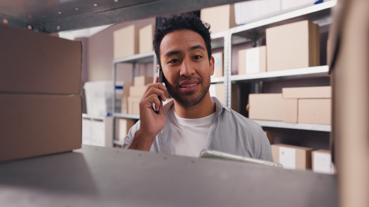 Happy Young Man Talking on Phone in Warehouse with Boxes