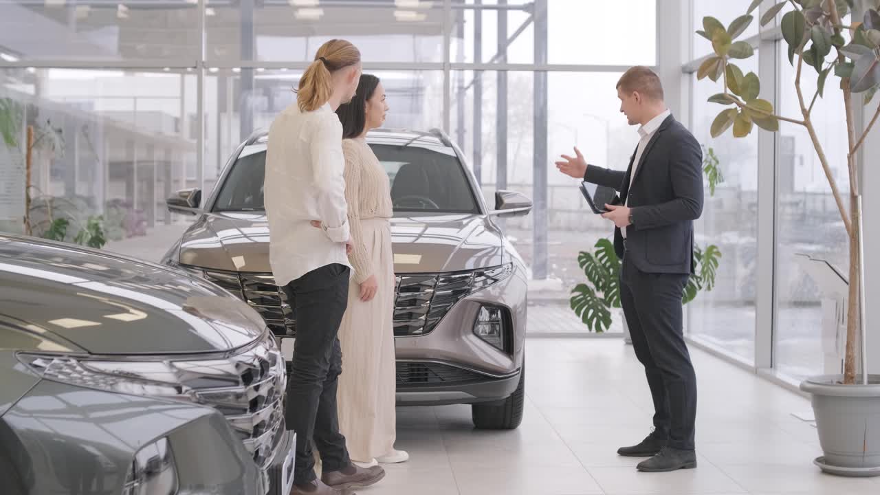 un joven vendedor de coches mostrando a una pareja de jóvenes un nuevo automóvil en el salón de concesionarios.