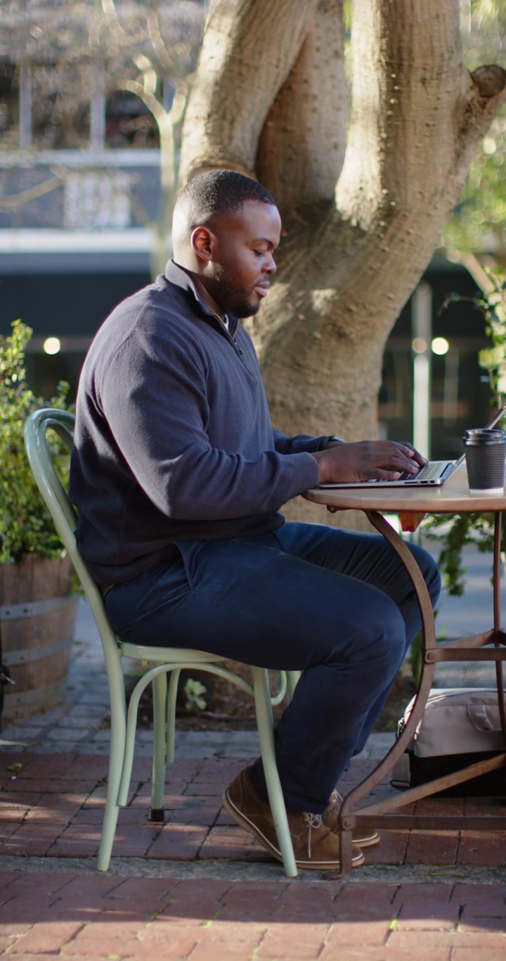 Vertical video: African American man typing on laptop working at café in sunlight sipping coffee