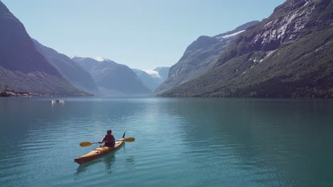 woman kayaking turquoise lovatnet lake during sunny summer day - backward moving aerial close to water with kayak moving towards camera - beautiful norway mountain landscape with glacier in background