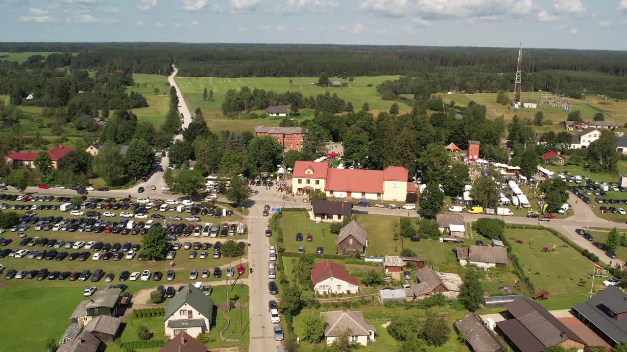 Aerial view of a small town hosting a lively outdoor fair with market stalls, parked cars, houses, and surrounding green fields under a sunny sky. Jaunpiebalga