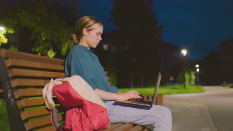 young woman sits on outdoor bench at night, using laptop under soft illumination in tranquil park setting, red backpack beside her, with bicycle and pathway lights in background