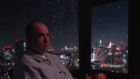 a man sitting looking out the window and rain in the city at night