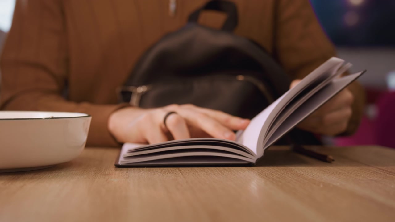Primo piano di una persona che sfoglia le pagine di un quaderno aperto su un tavolo di legno con illuminazione soffusa. La persona, che indossa abiti casual e porta una borsa nera, interagisce naturalmente mentre gira ogni pagina