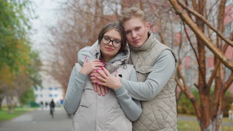 beautiful moment of lovely couple warmly holding each other as man gently rests head on her head in cozy outdoor scene with bare trees and blur background featuring people walking by in distance