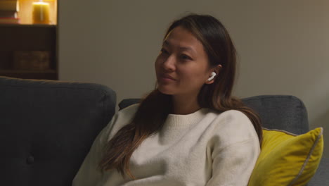 woman spending evening at home on sofa wearing wireless earbuds and streaming music or podcast