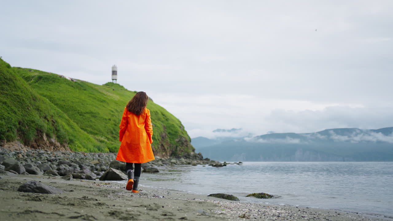 Woman in an orange raincoat walking on a beach by the ocean.