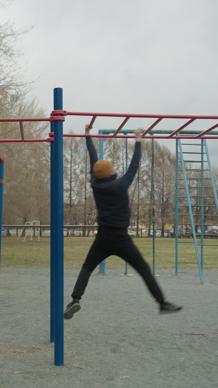 A coach leaps up to grab a red iron bar, then moves with controlled precision as he swings across it, in the background, a boy plays football on a field with park cars seen from a distance