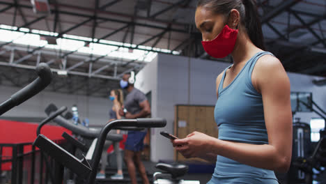 Frau-Mit-Gemischter-Rasse-Trägt-Gesichtsmaske-Und-Benutzt-Smartphone-Im-Fitnessstudio