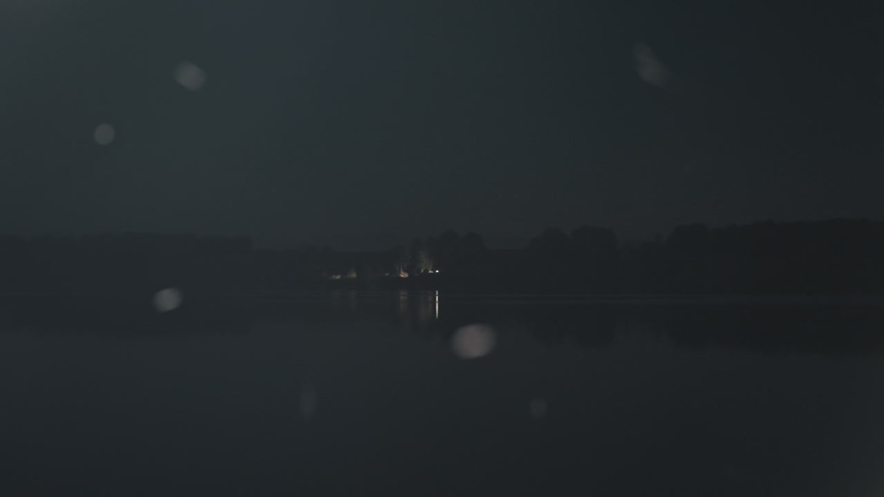 Nattlig campingscene ved innsjø med fjerne lys, myke snøfnugg som driver over rolig vann, gløden fra en ensom hytte på motsatt bredd, silhuetter av trær, svake refleksjoner, en camper som holder en lykt, og en fisker.