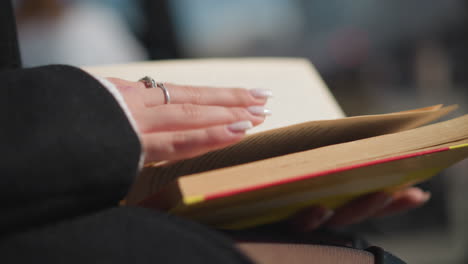 close-up of a woman with manicured nails gently flipping through the pages of a book, revealing a vibrant edge with sunlight reflecting off her hand and the surrounding soft background