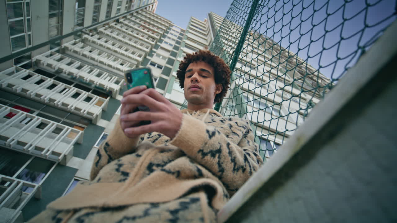Gadehipster bruger smartphone ved byhegn, fokuseret på skærmen. Latinamerikansk fyr.