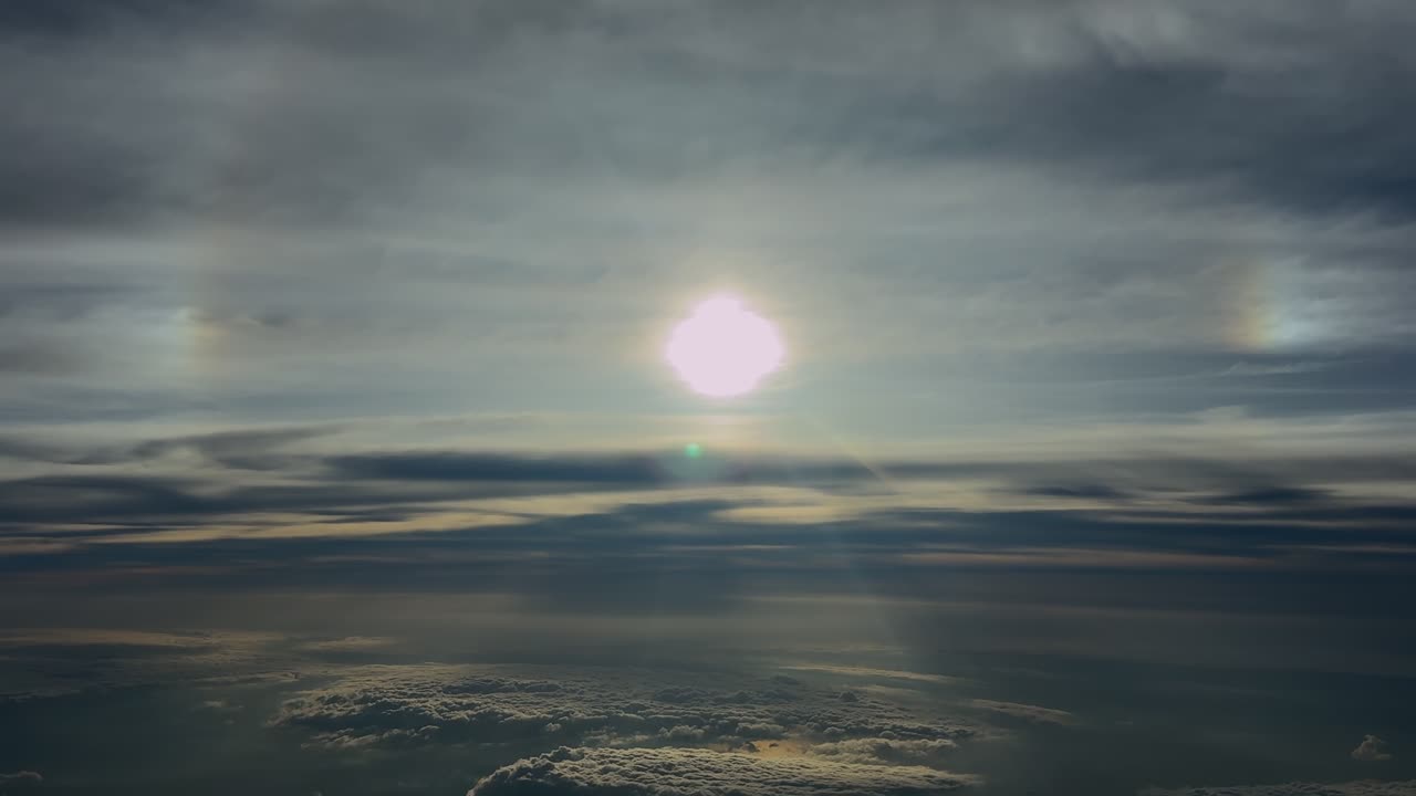 Et luftfoto fra et cockpit under en stratosfærisk flyvning med en bleg, glødende sol og dens glorie på en dramatisk himmel med lag af æteriske skyer.
