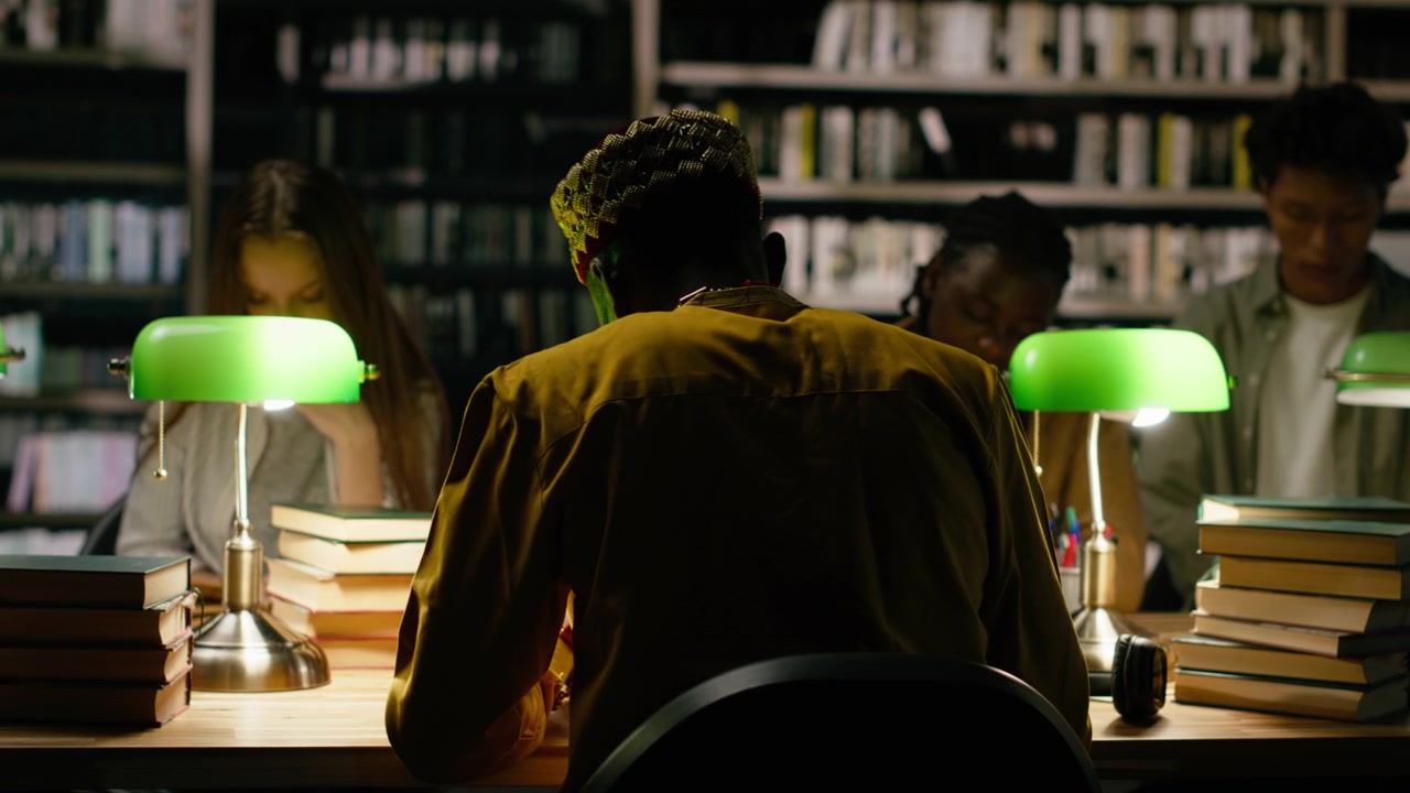 Vista da dietro di un giovane adulto con le cuffie che studia a un tavolo scuro della biblioteca.