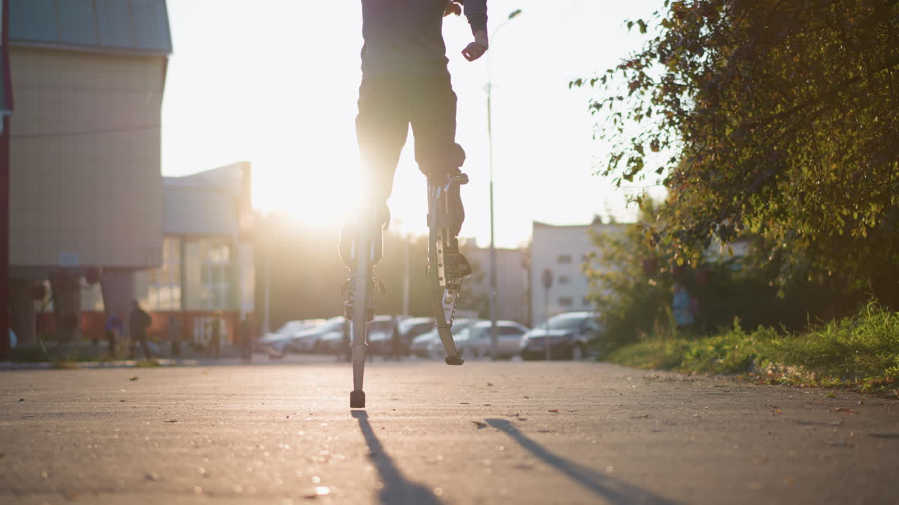 회색 후디와 어두운 회색 바지를 입은 남자의 뒷 장면은 해가 지는 동안 집으로 둘러싸인 주거 지역의 포장 된 도로에서 봄의 기둥에 뛰어 다니며 역동적인 움직임을 보여줍니다.