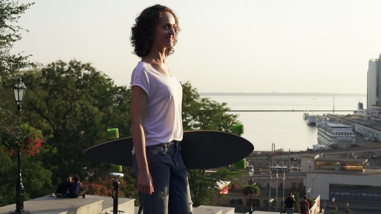 Young attractive girl walking holding her longboard in the city street during sunrise, slow motion shot. Beautiful city harbor