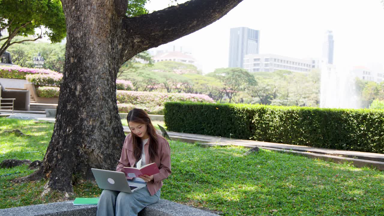 A young woman studies outdoors in a sunny park using a laptop and textbook