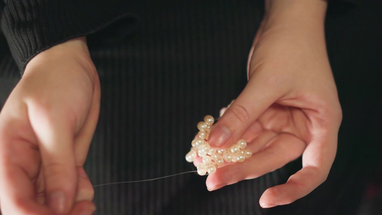 Closeup hands threading pearl beads, crafting quiet ritual as artisan forms delicate charm for foresight soft warm lighting, black sweater backdrop, focused fingers, slow deliberate movements, subtle