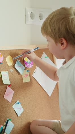 blond preschooler draws with pen on piece of paper slow motion. toddler boy learns to write and draw sitting on desk and playing with cork board in light room side view