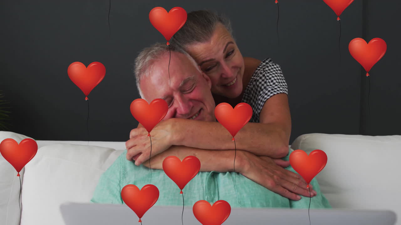 Casal idoso abraçado no sofá em cena de campanha de saúde, apresentando laptop e balões de coração flutuantes