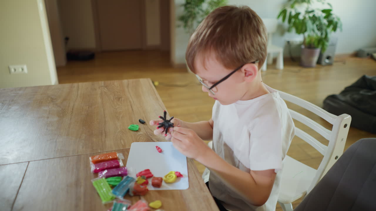 Boy with glasses sitting at wooden table molding colorful clay pieces into shapes with focus and creativity, engaging in artistic activity, developing imagination at home