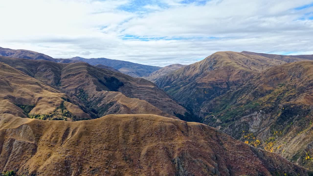 Drone ascends to reveal Macetown Road leading into Arrowtown, surrounded by autumn colors under cloudy skies with hints of blue