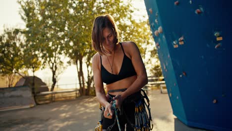 Portrait-of-a-blonde-girl-with-a-bob-hairstyle-who-ties-a-special-knot-on-her-climbing-equipment-to-create-insurance-and-looks-at-the-camera-near-a-blue-climbing-wall-on-a-sunny-summer-day