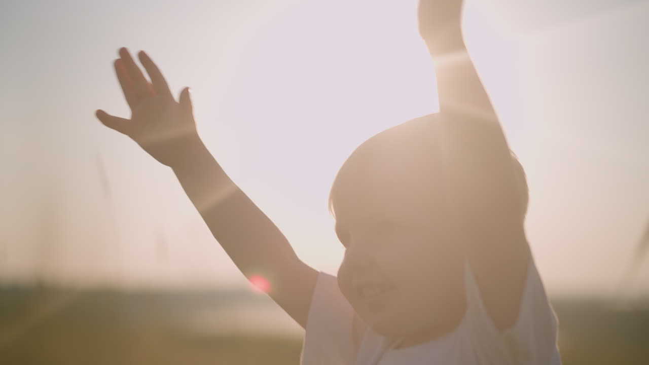 A joyful little boy in a white shirt walks through a sunny grass field with his hands raised, his face beaming with happiness, capturing a pure moment of childhood joy and wonder in nature