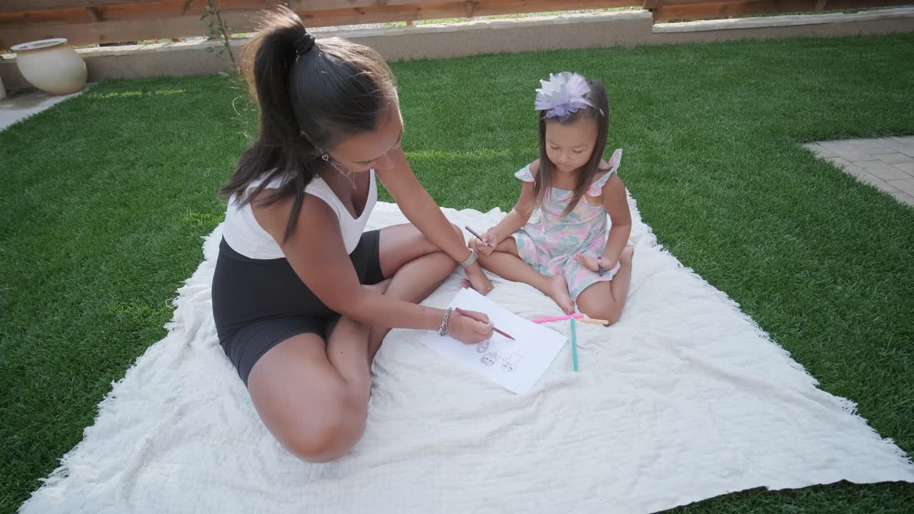 엄마와 어린 딸이 뒷마당에서 담요 위에 앉아 함께 그림을 그리고 있습니다. 야외 장면은 평화로운 가족 순간을 포착하여 창의성과 유대를 촉진합니다.