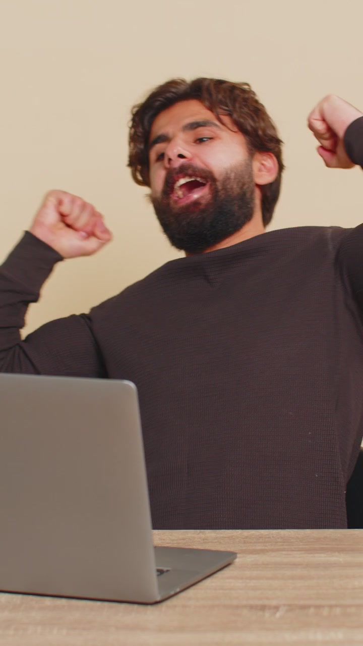 Hombre feliz sentado a la mesa usando una laptop, ganando la lotería y celebrando el éxito sobre un fondo beige.