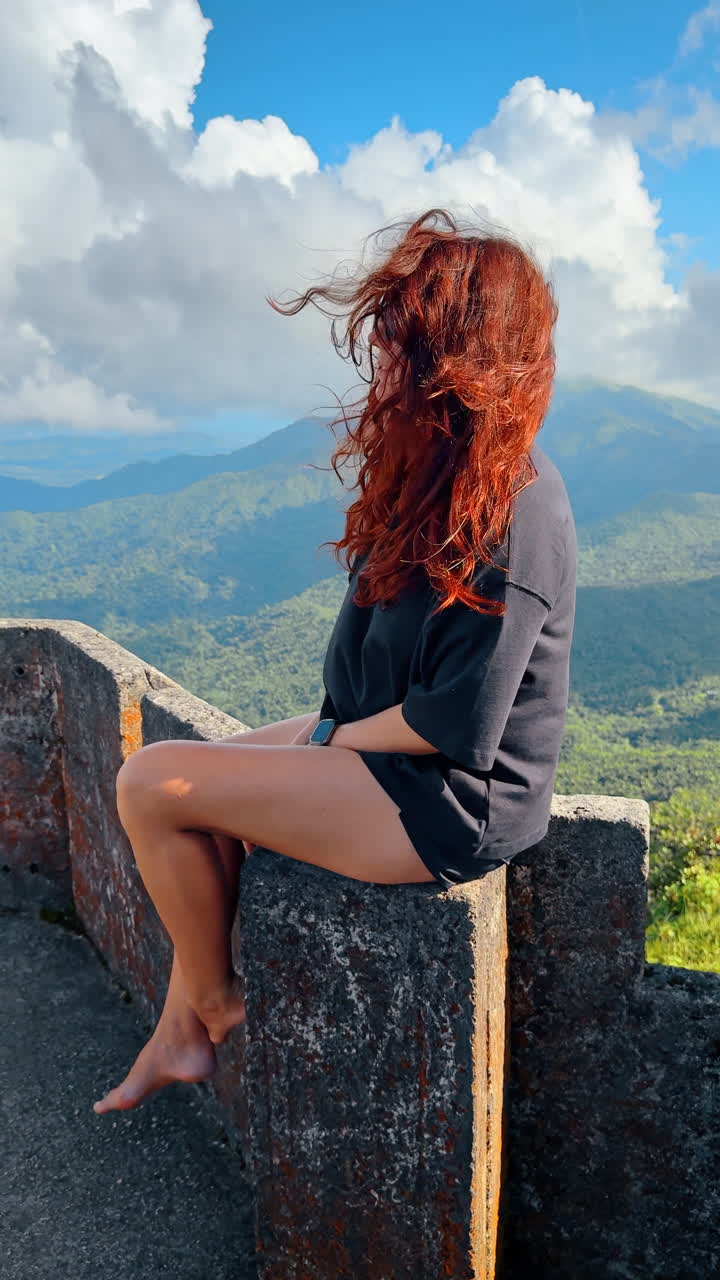 Red-haired girl wearing black t-shirt sits on the stone bastille. Picturesque mountains at backdrop. Vertical video