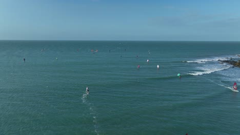 aerial dolly in flyover showing kitesurfers gliding across turquoise waves near rocky coastline in jericoacoara, brazil, under a clear blue sky
