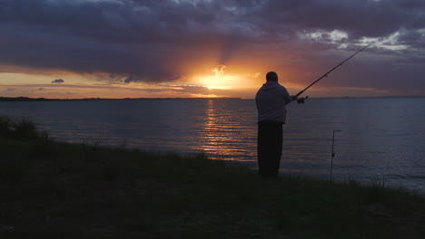 a lone fisherman casting his line from the banks of calm waters with an orange sun setting in the evening