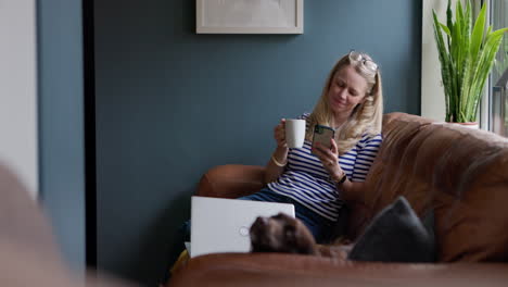 woman relaxes on couch with coffee, phone, dog, and laptop