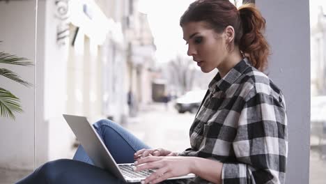 concentrated long haired beautiful girl working on laptop in light workspace. side view. blurred background