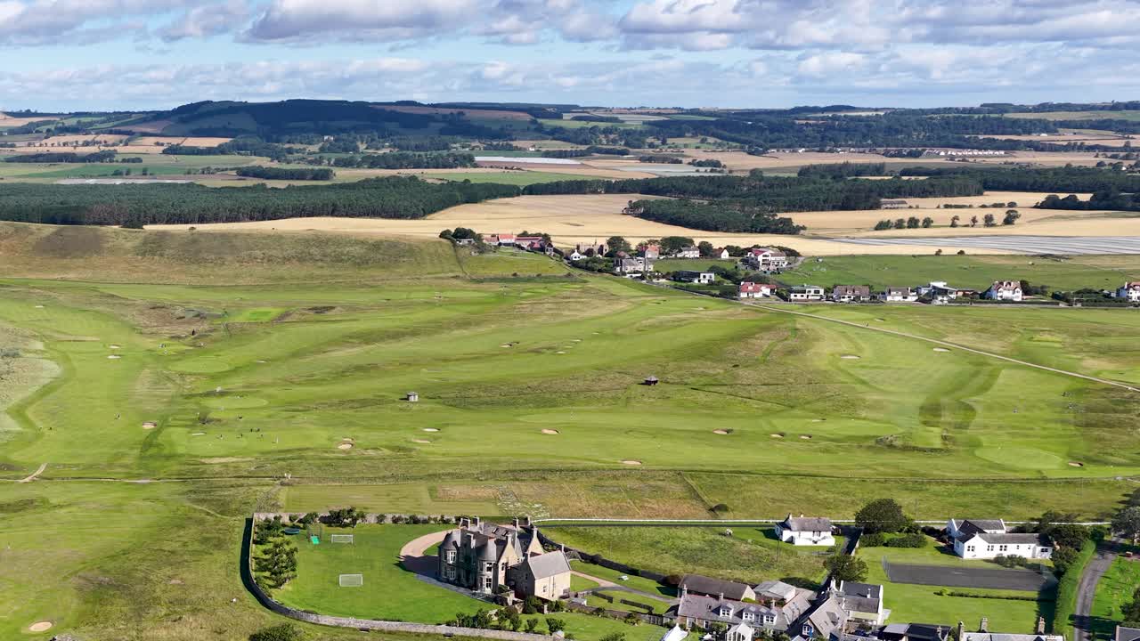 Drone pans across lush golf course, village, and coastal cliffs in bright daylight, wide perspective