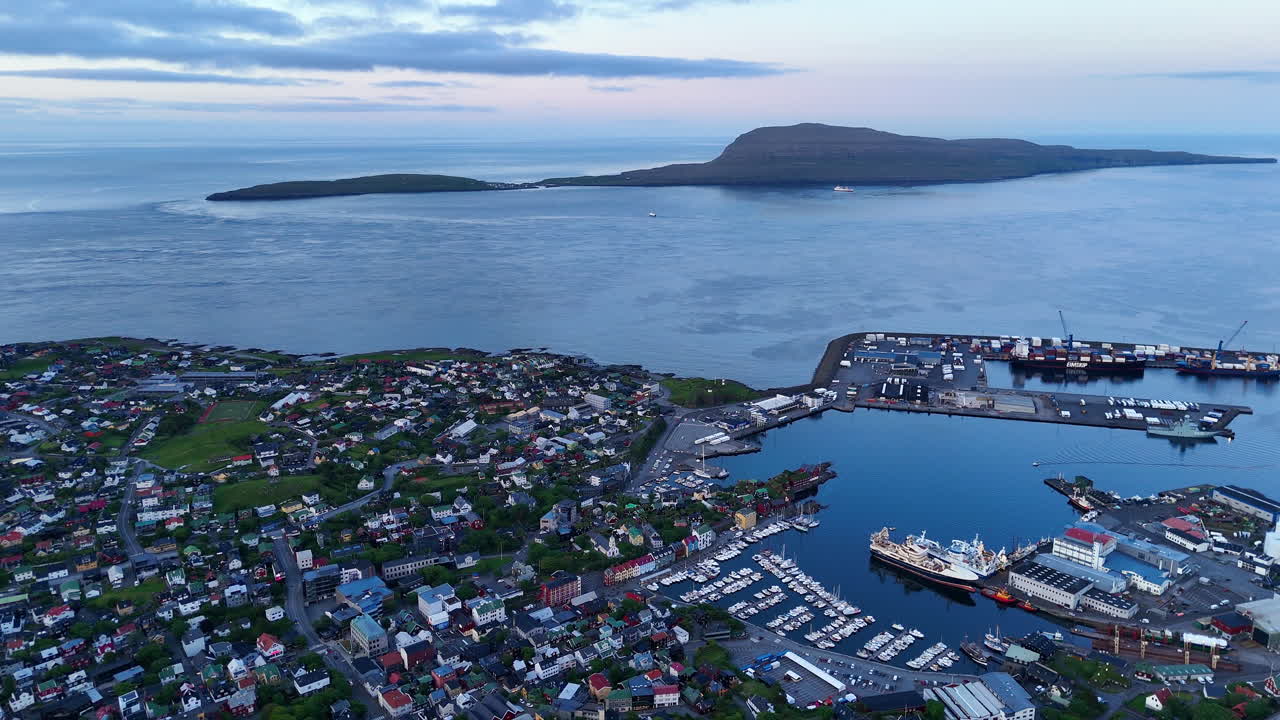 Cinematic aerial view of colorful village houses along a fjord in the Faroe Islands, surrounded by dramatic green mountains, black sand beach, and misty coastal scenery