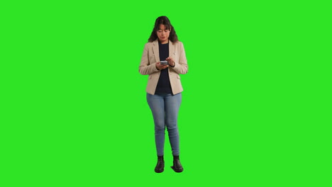 Full-Length-Studio-Portrait-Of-Female-Teacher-Or-Businesswoman-Standing-Against-Green-Screen-Using-Mobile-Phone-To-Message-Or-Browse-Online