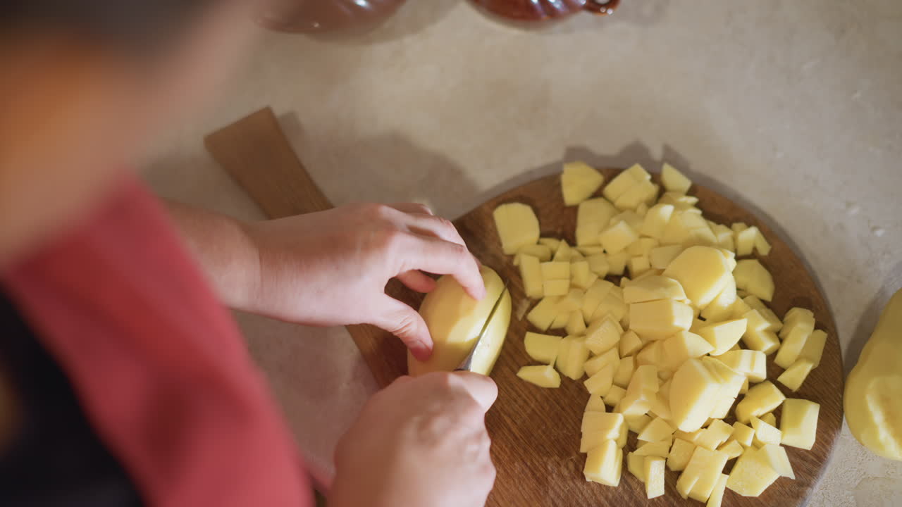 Manos de niño en cocina casera cortando patata, joven aprendiz corta cuidadosamente patata en tabla de madera, cálida luz matutina, primer plano lento, lección de cocina familiar, técnica para principiantes, receta acogedora de fin de semana.